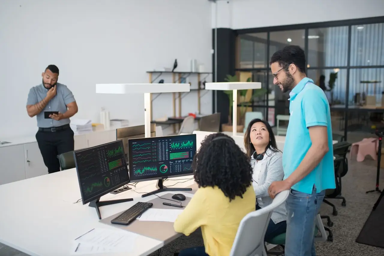 An open-plan office floor, colleagues talking beside a bank of monitors.