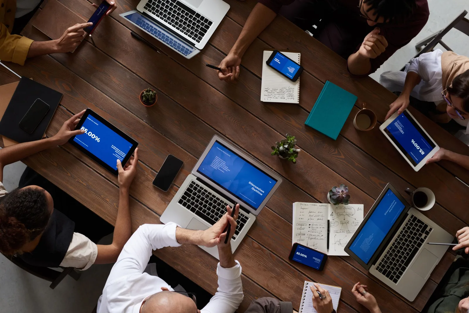 Overhead view of a team around a wooden table, laptops and tablets showing blue slides beside handwritten notes